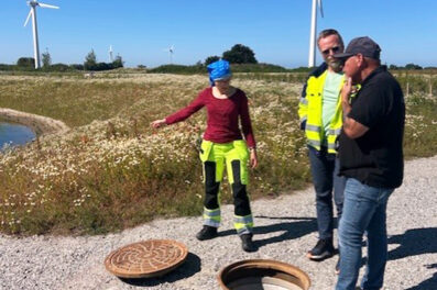 En kvinna och två män i grovarbetskläder står vid en äng och tittar ner i en öppen brunn på vägen. Brunnslocket ligger vid sidan av öppningen.