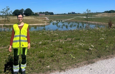 Leende kvinna i varselkläder står på en gång framför en gräsmatta. I bakgrunden in liten damm. Himlen är blå.