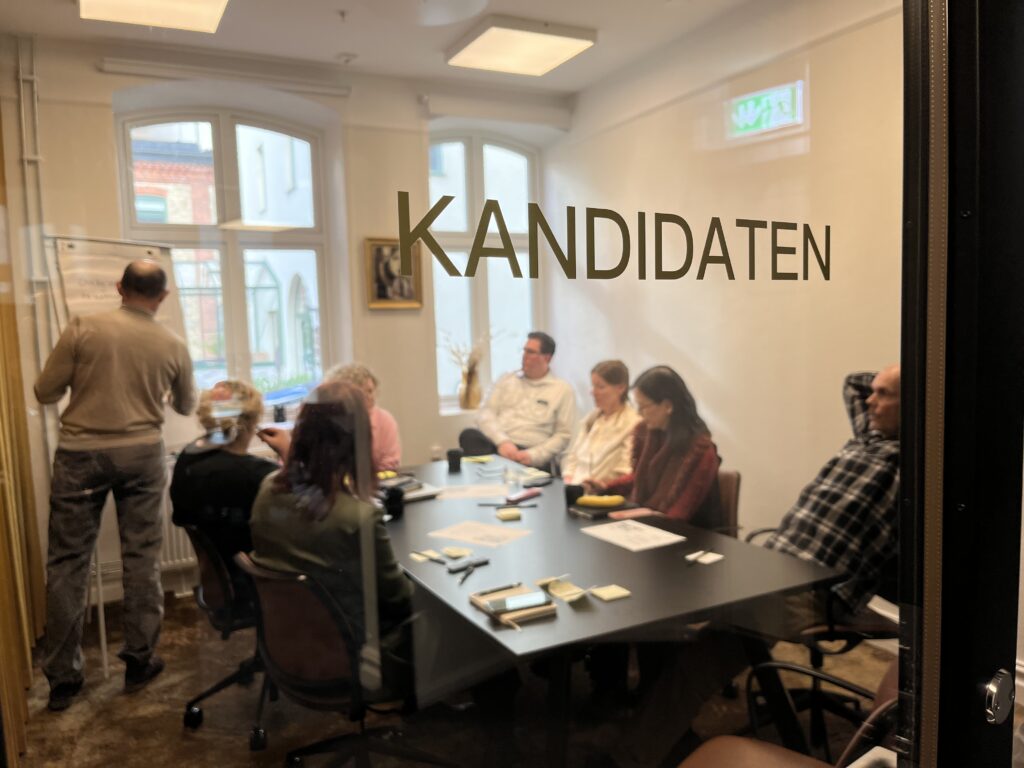 Människor runt ett konferensrum. De syns genom en glasruta med texten KANDIDATEN på.