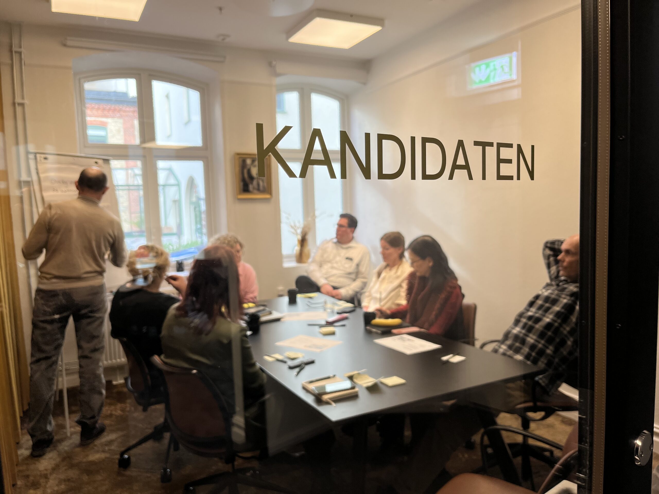 Människor runt ett konferensbord. De syns genom en glasruta med texten KANDIDATEN på.