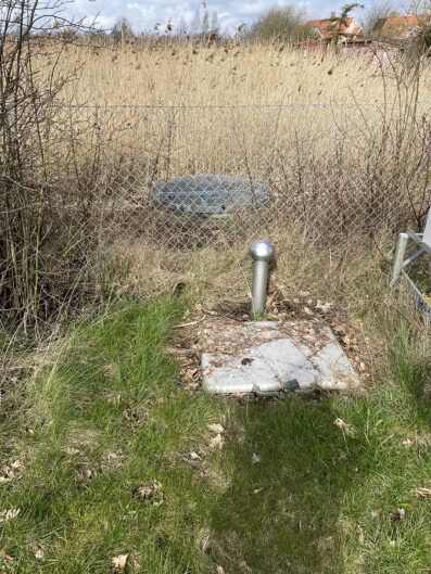 Rör som sticker upp ur marken vid en platta. Omgivet av gräs och ett stängsel bakom.