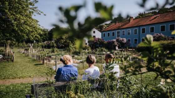 Tre personer bakifrån sittandes på en bänk. Grönt och lummigt, ett långt och blått tvåvåningshus i ena kanten.