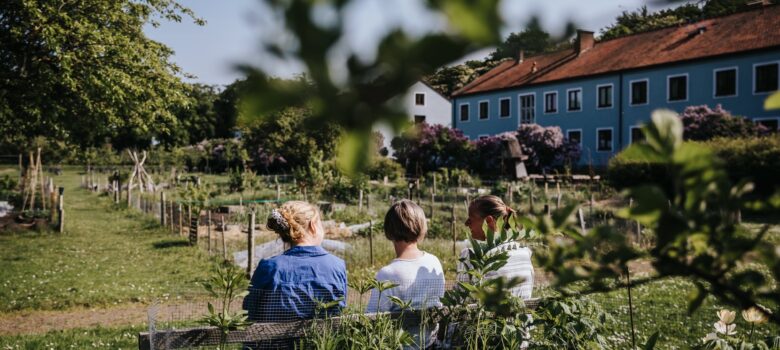 Tre personer bakifrån sittandes på en bänk. Grönt och lummigt, ett långt och blått tvåvåningshus i ena kanten.